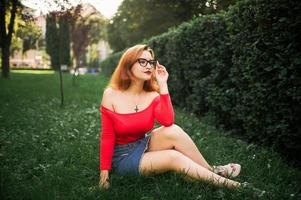 attraente donna dai capelli rossi in occhiali, indossare su camicetta rossa e gonna di jeans in posa al parco verde. foto