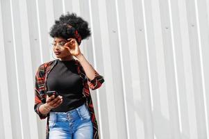 donna africana con capelli afro, in pantaloncini di jeans e occhiali posati contro la parete in acciaio bianco con il telefono cellulare in mano. foto