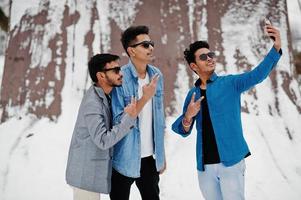 un gruppo di tre giovani indiani casuali in occhiali da sole posati in una giornata invernale e facendo selfie sul telefono. foto