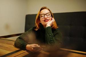 giovane e bella donna dai capelli rossi allegra con gli occhiali, indossa un maglione di lana caldo verde, seduto al bar e usando il suo telefono cellulare. foto