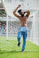 uomo a torso nudo muscoloso afroamericano bello sexy in tuta di jeans posato all'erba verde contro i cancelli di calcio del campo dello stadio. ritratto di uomo nero alla moda. foto