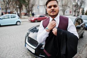 elegante uomo d'affari indiano in abbigliamento formale legato la cravatta e in piedi contro un'auto nera d'affari sulla strada della città. foto