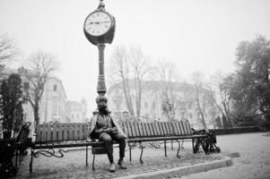 elegante modello uomo afroamericano in cappotto grigio, cravatta giacca e cappello rosso posato in panchina contro un grande orologio. foto in bianco e nero.