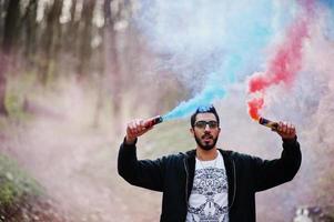 l'uomo arabo di street style con gli occhiali tiene il bagliore della mano con una bomba fumogena rossa e blu. foto