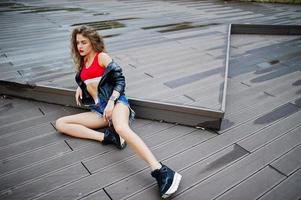 ragazza modello riccia sexy in top rosso, pantaloncini di jeans, giacca di pelle e scarpe da ginnastica poste sul pavimento. foto