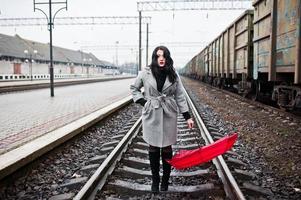 ragazza bruna in cappotto grigio con ombrello rosso nella stazione ferroviaria. foto