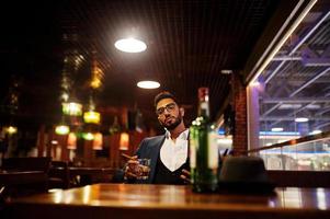bell'uomo arabo ben vestito con bicchiere di whisky e sigaro tenere il cellulare, posato al pub. foto
