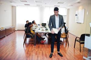 volto di un bell'uomo d'affari asiatico, tenendo il laptop sullo sfondo della riunione del team multirazziale della gente d'affari, seduto al tavolo dell'ufficio. foto