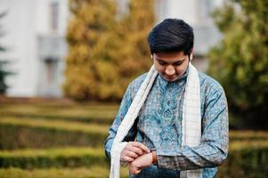 l'uomo indiano indossa abiti tradizionali con una sciarpa bianca posata all'aperto contro i cespugli verdi al parco, guardando i suoi orologi. foto