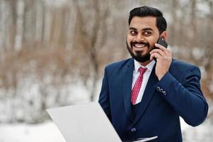 elegante uomo d'affari con barba indiana in giacca e cravatta posato in una giornata invernale all'aperto con un laptop a portata di mano, parlando sul telefono cellulare. foto