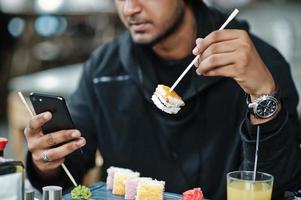 foto ravvicinata di un giovane asiatico casual ed elegante al bar che mangia sushi e guarda il telefono cellulare.