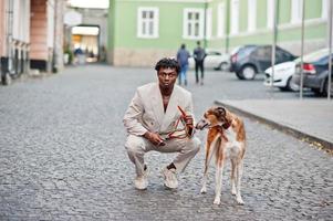 elegante uomo afro in abito beige vecchia scuola con cane borzoi russo. giovane maschio africano alla moda in giacca casual sul torso nudo. foto
