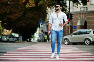 elegante uomo arabo alto modello in camicia bianca, jeans e occhiali da sole posato in una strada della città. barba attraente ragazzo arabo con una tazza di caffè che cammina sul passaggio pedonale o sul passaggio pedonale. foto