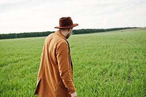 uomo alla moda con gli occhiali, giacca marrone e cappello in posa sul campo verde. foto