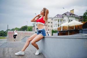 ragazza modello riccia sexy in top rosso, pantaloncini di jeans, occhiali da vista e scarpe da ginnastica poste allo skatepark. foto