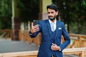 uomo d'affari indiano con barba elegante con bindi sulla fronte, indossare un abito blu posato all'aperto e fare selfie al telefono cellulare. foto