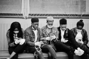 gruppo di amici adolescenti africani seduti con i telefoni, che indossano maschere mediche proteggono da infezioni e malattie quarantena del virus del coronavirus. foto