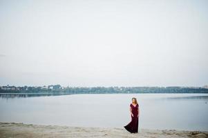 bionda sensuale donna a piedi nudi in abito rosso marsala in posa contro il lago sulla sabbia. foto