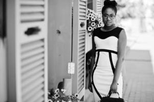ragazza afroamericana con occhiali, abito bianco e nero e borsa in posa contro finestre viola. foto