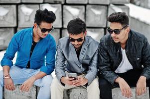 un gruppo di tre giovani indiani casuali in occhiali da sole posati contro blocchi di pietra e guardando il telefono cellulare. foto