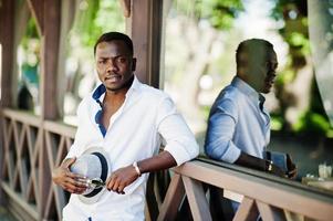 elegante uomo afroamericano in camicia bianca e pantaloni colorati con cappello in posa all'aperto. ragazzo nero modello alla moda. foto