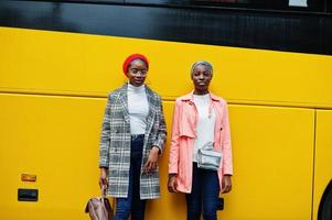 due giovani donne musulmane africane moderne alla moda, attraenti, alte e snelle in hijab o turbante, sciarpa e cappotto poste contro l'autobus giallo. foto