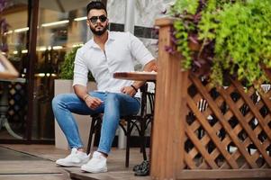 elegante uomo arabo alto modello in camicia bianca, jeans e occhiali da sole posato in una strada della città. barba attraente ragazzo arabo seduto sul caffè all'aperto. foto