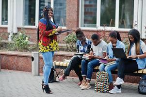 gruppo di cinque studenti universitari africani che trascorrono del tempo insieme nel campus nel cortile dell'università. amici afro neri che studiano al banco con articoli per la scuola, notebook portatili. foto