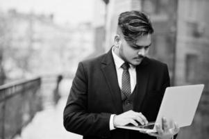 elegante uomo d'affari indiano in abbigliamento formale con laptop sulle mani in piedi contro le finestre nel business center. foto