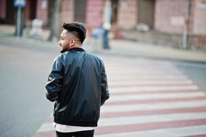 uomo alla moda con barba indiana in giacca di pelle nera che cammina sulle strisce pedonali. modello indiano posato all'aperto nelle strade della città. foto