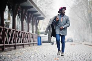 elegante modello uomo afroamericano in cappotto grigio, cravatta giacca e cappello rosso posato in strada con tempo nebbioso. foto