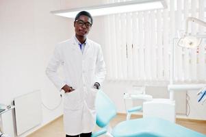 medico maschio afroamericano con gli occhiali in piedi vicino alla poltrona del dentista in clinica dentale. foto