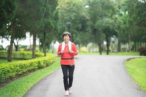 donna anziana asiatica atletica anni '60 sorridente e fare jogging. bella donna asiatica senior che corre al parco in una giornata di sole. corridore femminile che ascolta la musica mentre fa jogging. foto
