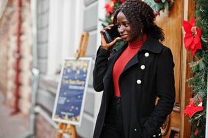 ritratto di una donna africana dai capelli ricci che indossa un cappotto nero alla moda e un dolcevita rosso in posa all'aperto vicino alla porta con decorazioni natalizie, capodanno. parlare per telefono. foto