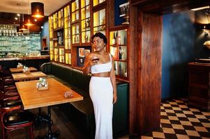 donna afroamericana, acconciatura retrò in abito bianco al ristorante con bicchiere di vino. foto