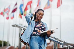 studentessa africana in posa con zaino e articoli per la scuola nel cortile dell'università, contro le bandiere di diversi paesi. foto