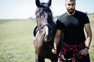 l'uomo arabo con barba alta indossa in nero con cavallo arabo. foto