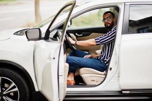 uomo arabo di successo indossa una camicia a righe e occhiali da sole posa al volante della sua suv bianca. uomini arabi alla moda in trasporto. foto