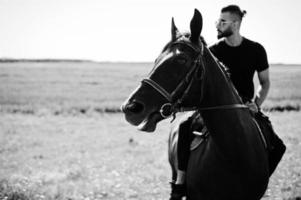 l'uomo arabo con barba alta indossa in nero e gli occhiali da sole cavalcano un cavallo arabo. foto