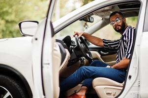 uomo arabo di successo indossa una camicia a righe e occhiali da sole posa al volante della sua suv bianca. uomini arabi alla moda in trasporto. foto