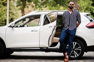 uomo arabo di successo indossa una camicia a righe e occhiali da sole posa vicino alla sua auto suv bianca. uomini arabi alla moda in trasporto. foto