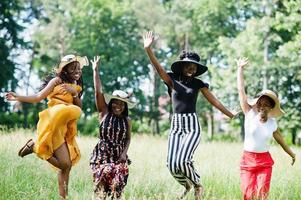 un gruppo di quattro bellissime donne afroamericane indossano un cappello estivo che salta all'erba verde nel parco.