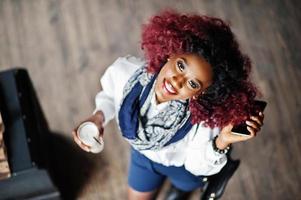 attraente ragazza riccia afroamericana in camicetta bianca e pantaloncini blu poste al bar con latte e telefono cellulare a portata di mano. vista dall'alto. foto