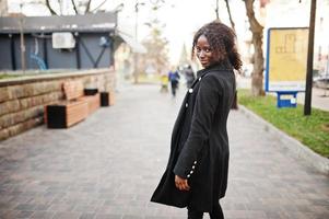 ritratto di una donna africana dai capelli ricci che indossa un cappotto nero alla moda e un dolcevita rosso in posa all'aperto. foto