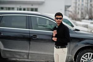 casual giovane indiano in camicia nera e occhiali da sole in posa contro l'auto suv. foto