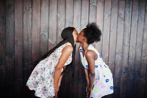 due amiche nere africane in abiti estivi poste su uno sfondo di legno scuro e si baciano. foto