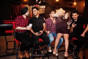 gruppo di amici indiani seduti al lounge bar, divertendosi e riposando, fumando narghilè e guardando sui telefoni cellulari. foto