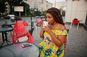 carina ragazza afroamericana di piccola altezza con i dreadlocks, indossa un abito giallo colorato, seduta al bar all'aperto su una sedia rossa e bevendo caffè. foto