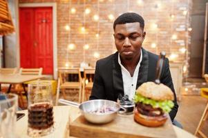 mordersi il labbro. rispettabile giovane afroamericano in abito nero seduto al ristorante con gustoso doppio hamburger e bibita gassata. foto