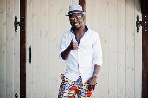 elegante uomo afroamericano in camicia bianca e pantaloni colorati con cappello e occhiali in posa all'aperto. ragazzo nero modello alla moda. foto